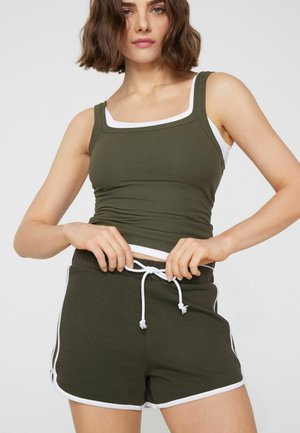 Frau trägt ein olivgrünes, geripptes Tanktop und passende Shorts mit weißem Rand, zieht die Kordel an den Shorts fest.