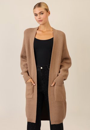 Langer, offen gestalteter Cardigan aus beigem Rippstrickstoff, mit zwei Fronttaschen und oversized Ärmeln, getragen über einem schwarzen Oberteil und Jeans.