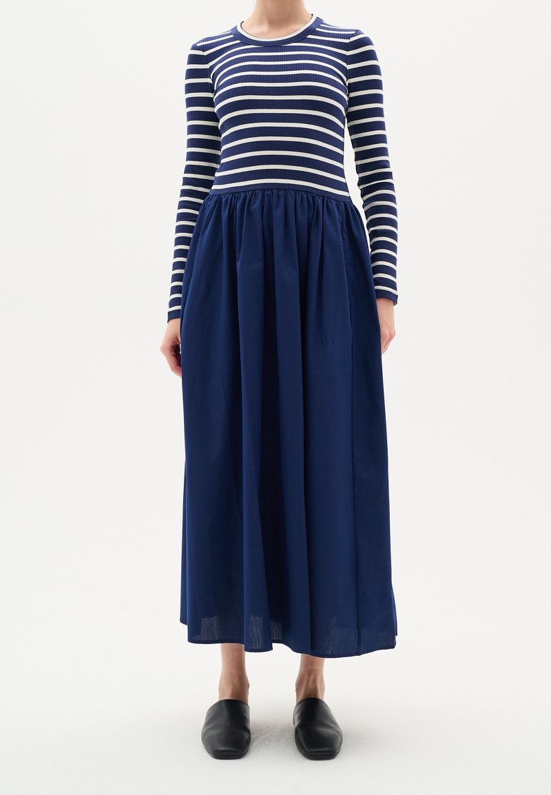 Robe à manches longues rayée avec un haut côtelé ajusté bleu marine et blanc, se déployant en une jupe fluide et ample bleu marine, associée à des chaussures noires sans lacets.