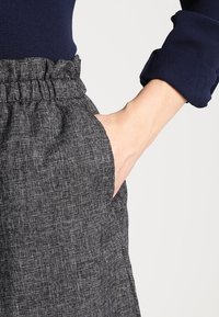 Ruka v kapse šedých texturovaných kalhot s vysokým pasem a elastickým pasem, které jsou kombinovány s námořnickou modrou halenkou s dlouhými rukávy a ohrnutými manžetami.