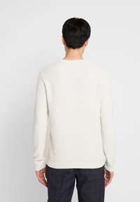 Pull en maille beige clair avec un motif texturé, col rond et poignets côtelés, associé à un pantalon à carreaux sombre.