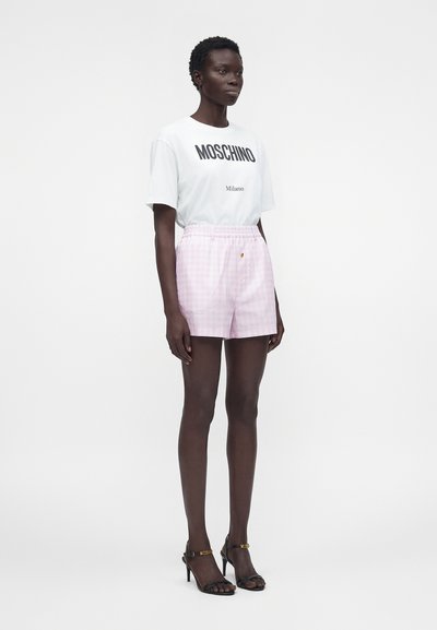 Femme debout portant un t-shirt blanc Moschino Milano, un short rose à carreaux avec des boutons et des sandales à talons hauts noires avec logo.