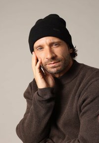 Zwarte beanie muts met geribbelde textuur en een klein label, gecombineerd met een bruine geribbelde trui. Model poseert met hand op gezicht.