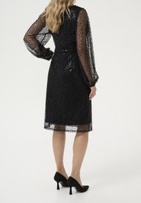 Robe en dentelle noire avec des manches longues et transparentes, présentant un motif texturé et une coupe jusqu'aux genoux. Associée à des chaussures à talons hauts noires.