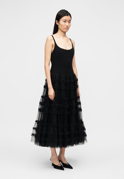 Μαύρο φόρεμα με εφαρμοστό μπούστο και στρωμένο τούλινο φούστα με βολάν στο τελείωμα. Το μοντέλο φορά μαύρες μυτερές μπαλαρίνες. Ομαλή υφή συνολικά.