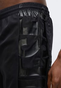 Μαύρο αθλητικό σορτς με γυαλιστερή, υφή πλευρική επιφάνεια με γεωμετρικές οπές. Ελαστική ζώνη μέσης για ασφαλή εφαρμογή και ευελιξία.