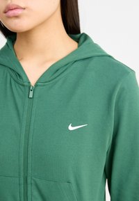 Zelená zipsová mikina vyrobená z mäkkého materiálu, s bielym logom Nike na ľavej hrudi a kapucňou so šnúrkou. Hladká textúra a neformálny dizajn.