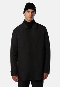 Black waterproof coat with a straight cut, large collar, side pockets, and buttoned cuff details. Worn with a black knit turtleneck and beanie.