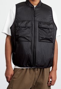Svart ärmlös väst av lättviktsmaterial, med hög krage, framsida med dragkedja och två bröstfickor. Strukturerad yta med logotyp.