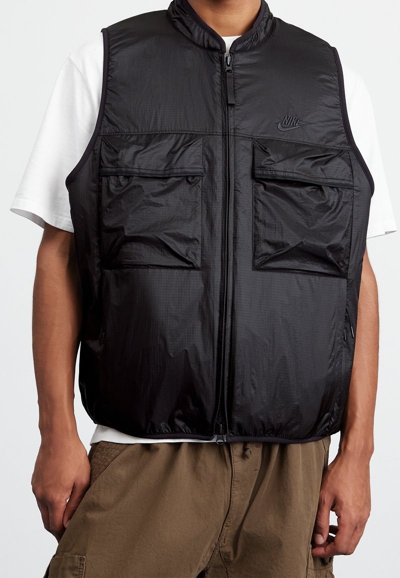 Svart ärmlös väst av lättviktsmaterial, med hög krage, framsida med dragkedja och två bröstfickor. Strukturerad yta med logotyp.