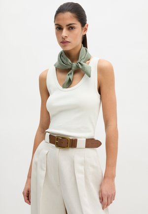 Vrouw draagt een wit mouwloos topje, beige broek met hoge taille en bruine riem, en een groen sjaaltje om haar hals geknoopt.