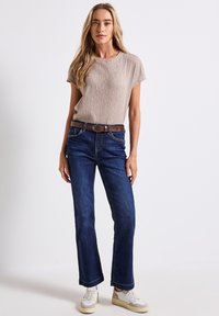 Haut beige texturé à manches courtes associé à un jean flare en denim bleu foncé. Une ceinture marron avec boucle en métal argenté et des baskets blanches complètent le look.