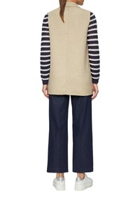 Beige ärmellose Tunika mit einem Kragen, kombiniert mit einem gestreiften Langarmshirt und dunkelblauen weit geschnittenen Hosen. Weiße Sneaker runden das Outfit ab.