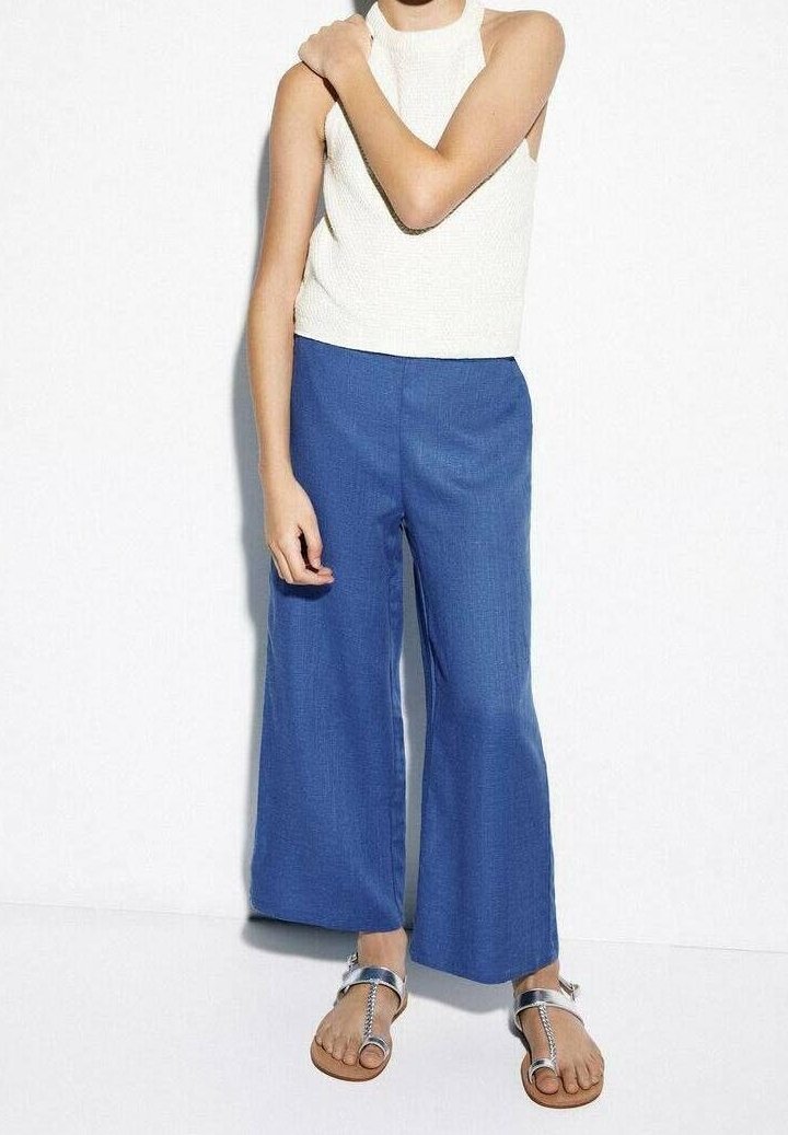 Femme portant un haut blanc sans manches, un pantalon large bleu et des sandales argentées, debout contre un mur blanc avec un bras croisé sur la poitrine.