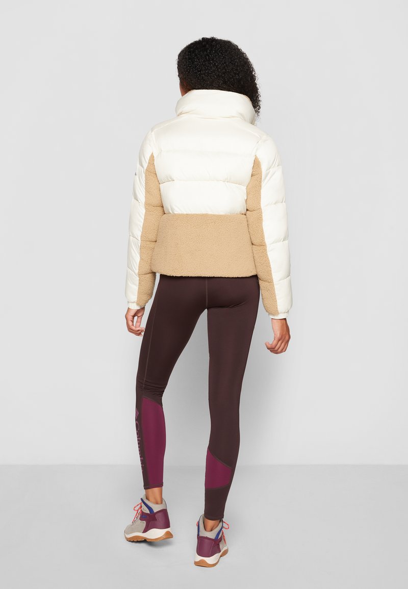 Giacca a vento con parte superiore crema e parte inferiore in pile color sabbia, dotata di collo alto e polsini elasticizzati, abbinata a leggings scuri e scarpe multicolori.