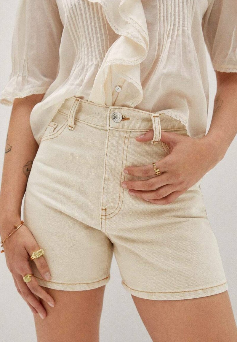 Shorts de denim beige con cintura alta, costuras en contraste, botón frontal y bolsillos. La mano del modelo descansa sobre la cintura.