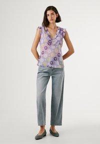 Lila blus med blommönster och volangärmar, knappar vid halsringningen, matchad med ljusgrå vida jeans och gråa platta skor.