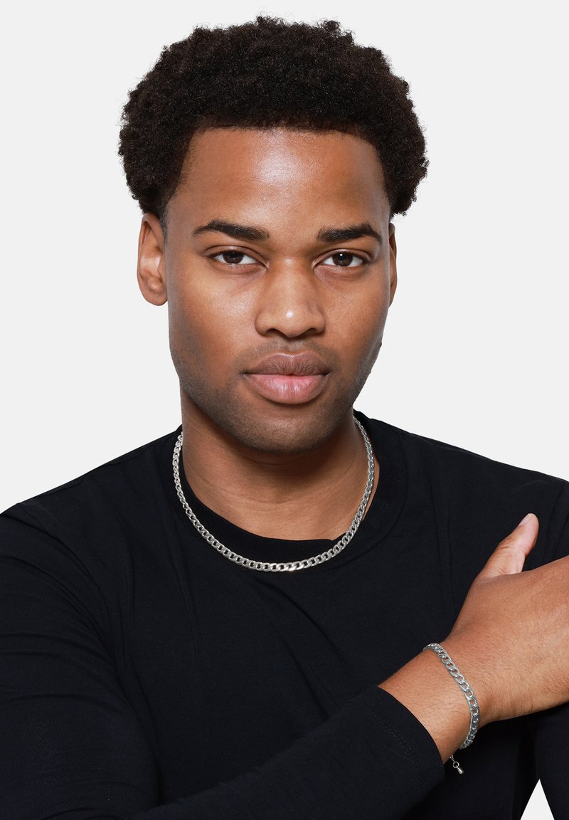 Sølvkjede og armbånd på en person som har på seg en langermet svart skjorte. Smykkene har et polert, sammenflettet design.