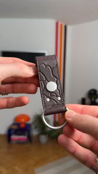 Pruunist nahast võtmehoidja, millel on reljeefne muster, hõbedane metallrõngas ja metallist brändiga ketas. Sile tekstuur ja dekoratiivsed detailid.