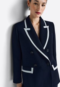 Blazer azul marino de doble botonadura con solapas blancas y detalles en los bolsillos. Hecho de tela suave, cuenta con botones negros, ajuste entallado y cuello con muesca.