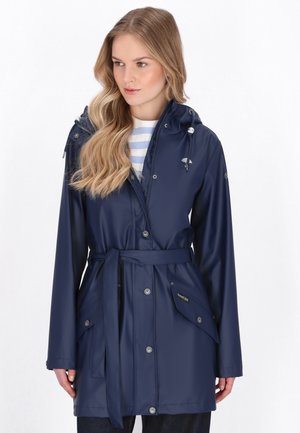 Kobieta w granatowym, zapinanym na pasek płaszczu przeciwdeszczowym z kapturem, mająca na sobie biało-niebieską pasiastą koszulę, stoi na tle jednolitego białego tła.