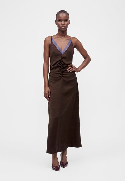 Femme portant une robe marron sans manches avec une bordure en dentelle bleue au niveau du décolleté et un détail froncé à l'avant, debout avec une main sur la hanche.