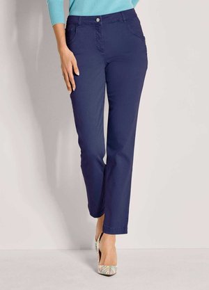 Vrouw in marineblauwe straight-leg broek en kleurrijke hoge hakken, staande tegen een effen lichte achtergrond.