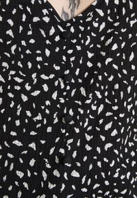 Tessuto nero con una superficie texture caratterizzato da un motivo astratto bianco. Dettagli notevoli includono due bottoni neri disposti verticalmente.
