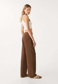 Hnědé kalhoty širokého střihu vyrobené z texturované látky, s vysokým pasem a přiléhavým střihem, doplněné o bílé crop top a sandály.