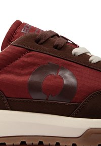 Gros plan du côté d'une basket marron et rouge avec des lacets bicolores et un logo flèche circulaire au-dessus de la semelle intermédiaire blanche.