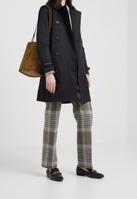 Osoba w czarnym dwurzędowym płaszczu, brązowych spódnicach w kratę, czarnych loafersach z złotym łańcuchem, niosąca dużą brązową torebkę z zamszu.