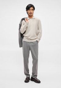 Polo pulover tricotat bej cu trei butoane, asociat cu pantaloni gri deschis, croiți, și pantofi maro închis. Jacheta gri pusă pe umăr.