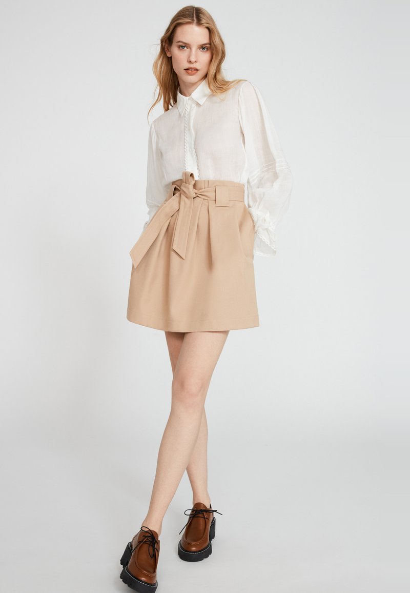 Beige Hochbundrock mit gebundenem Gürtel. Kombiniert mit einem weißen Langarmhemd. Das Model trägt braune Schnürschuhe mit dickem Absatz.