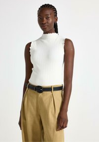 Top bianco a coste senza maniche con scollatura scallop, abbinato a pantaloni beige plissettati e una cintura nera con cuciture bianche.