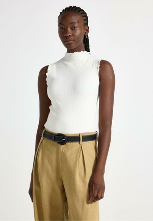 Vit randig ärmlös topp med en scallop-neckline, parat med beige veckade byxor och ett svart bälte med vit sömnad.