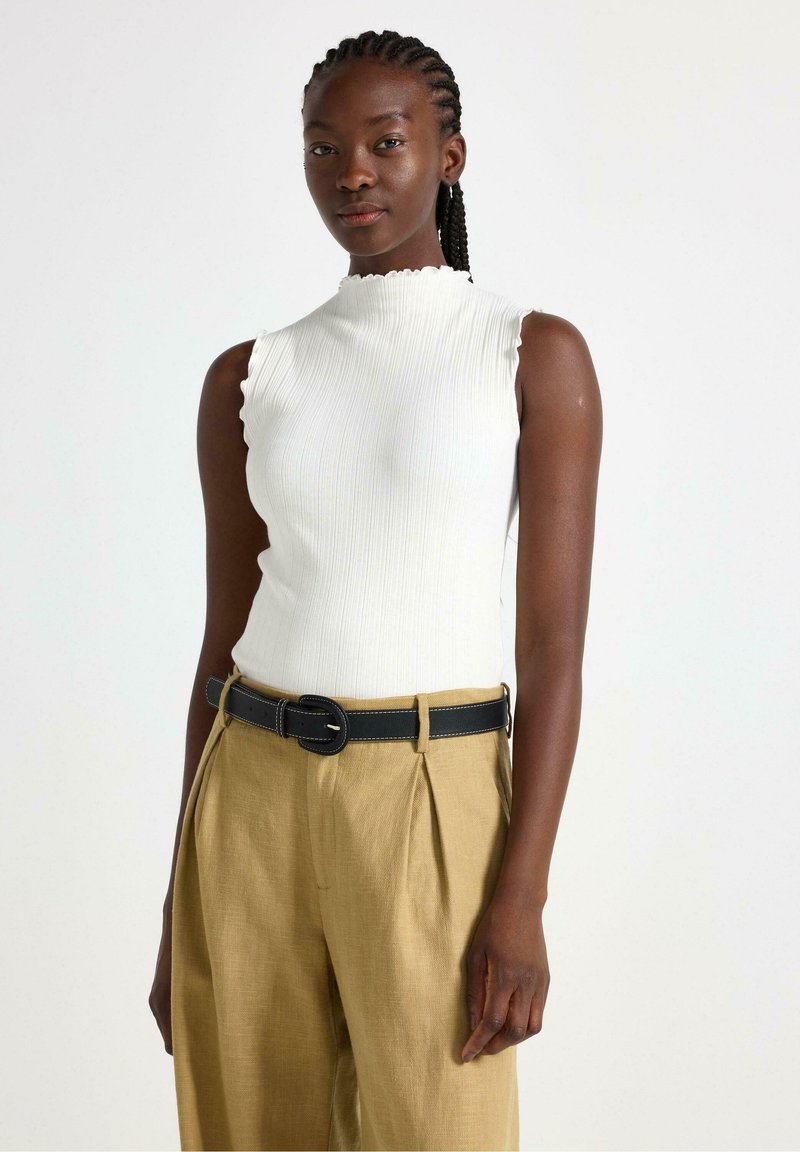Top bianco a coste senza maniche con scollatura scallop, abbinato a pantaloni beige plissettati e una cintura nera con cuciture bianche.
