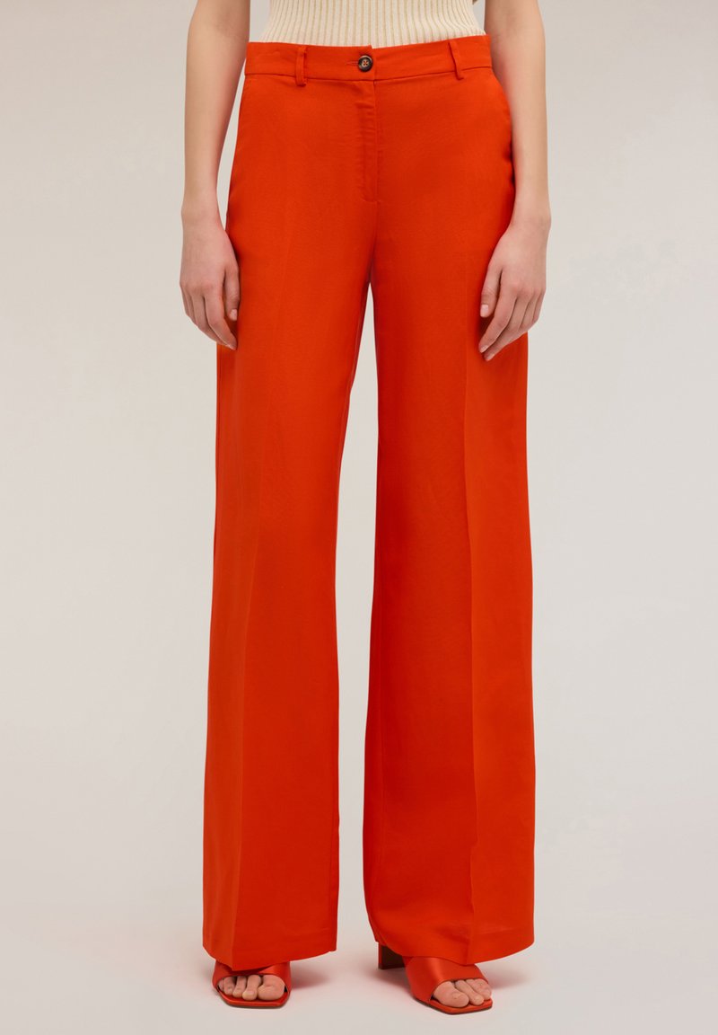 Pantaloni a gamba larga in tessuto rosso brillante, con chiusura a bottoni, vita alta e texture liscia. Abbinati a sandali rossi coordinati.
