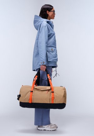 Borsa a sacca beige con base nera, manici arancioni e una tasca frontale. Il modello indossa una giacca con cappuccio azzurro chiaro e pantaloni abbinati.