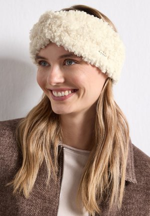 Femme souriante portant un bandeau duveteux de couleur crème et une veste marron, regardant légèrement vers la droite.