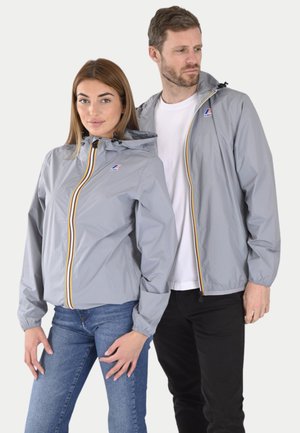 Grå vindjakker med hette, med glidelås foran og gule og røde aksentstriper, båret av to modeller med jeans.