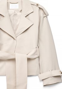 Gabardina beige con cuello de solapa ancho, cinturón anudado a la cintura, hombreras con botones y correas ajustables en los puños de las mangas largas.