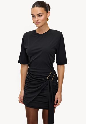 Femme avec les cheveux plaqués en arrière, portant une mini-robe noire à fronces avec des manches courtes et des boucles d'oreilles en forme de hoops dorées, sur fond blanc.