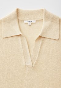 Polo en maille beige avec un col en V ouvert et une étiquette indiquant OPUS taille 36 à l'arrière à l'intérieur de l'encolure.