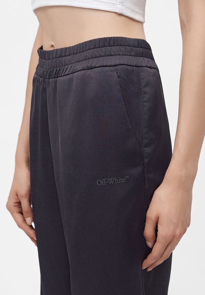 Personne portant un pantalon de survêtement noir Off-White avec une taille élastique et un haut court blanc, montrant le ventre et une position de main détendue.
