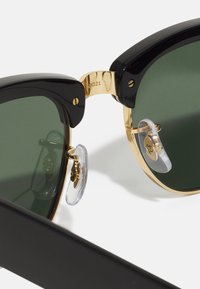 Ochelari de soare retro negri cu lentile verzi, accente metalice aurii și tampoane transparente pentru nas. Cadre groase cu un design lat, evidențiind un detaliu al balamalei.