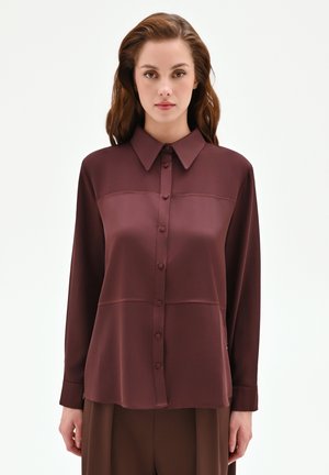 Donna con capelli castani ondulati che indossa una camicetta bordeaux a maniche lunghe con bottoni e pantaloni marroni, in piedi contro uno sfondo bianco semplice.