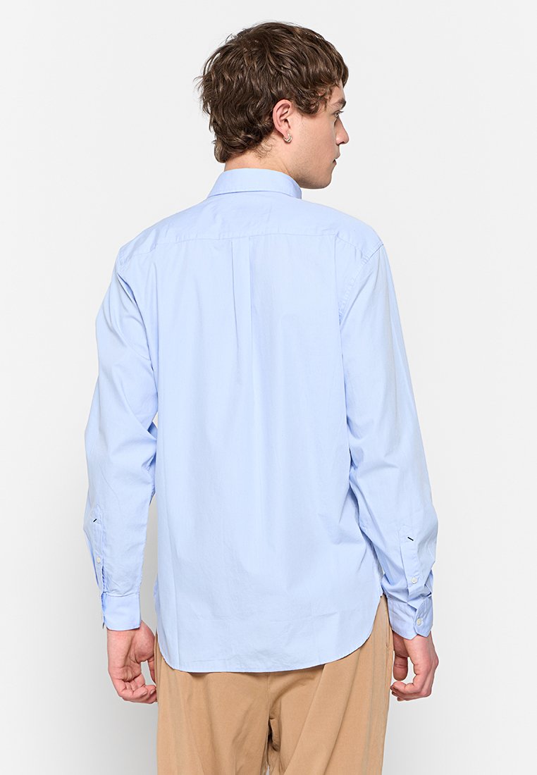 Junger Mann, der sich abwendet, trägt ein hellblaues Langarmhemd und beige Hosen, steht vor einem schlichten weißen Hintergrund.