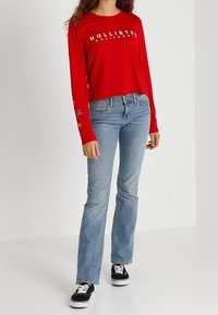 Röd långärmad skjorta med texten "HOLLISTER CALIFORNIA", tillsammans med ljusblå utsvängda jeans och svarta sneakers, som står mot en vit bakgrund.