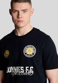 Svart t-shirt med kort ärm som har en cirkulär solroslogotyp och texten "SKINER ÖVER SKOTTLAND" tillsammans med "SUNNIES F.C." i vitt.