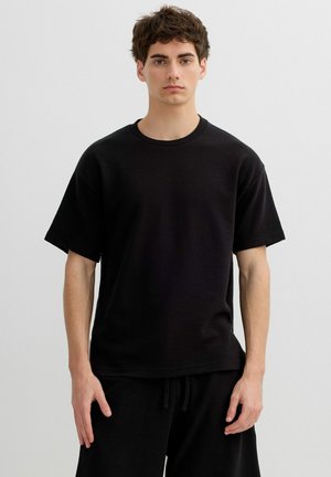 Joven con cabello corto y rizado, vistiendo una camiseta negra lisa y oversize junto con pantalones negros a juego, posando frente a un fondo gris.